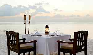 Ett dukat bord är placerat på en sandstrand nära vattnet med lyktor och facklor vid sidan. (AI-genererad text)