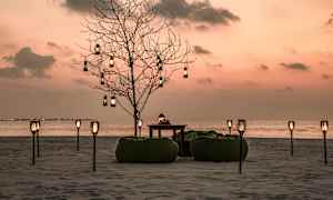 En strandmiljö under solnedgång med ett dekorerat träd, sittpuffar och bord tillsammans med avlånga lyktor. (AI-genererad text)