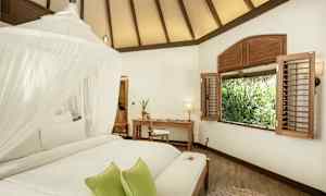 Ett ljust rum med ett vitt tak, en säng med myggnät och gröna kuddar, ett skrivbord och en stol vid ett fönster med utsikt över grönska. (AI-genererad text)