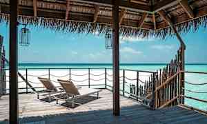 Två solstolar på en träbrygga med utsikt över en klarblå havskust under ett halmtak. (AI-genererad text)