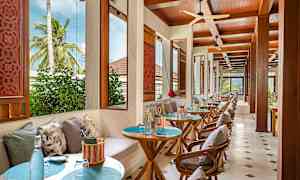 Bild av en restaurang med bord och stolar, dekorerade med kuddar och utsikt över växtlighet genom fönstren. (AI-genererad text)