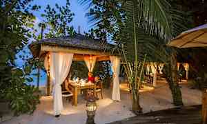 En upplyst uteplats med tak och vita gardiner, omgiven av grönska på en strand. (AI-genererad text)