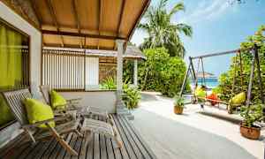 Ett uteområde med trästolar, en gunga och sand, omgiven av grönska och palmträd. (AI-genererad text)