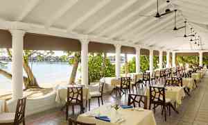 En restaurangs uteplats med dukade bord, omgiven av pelare och utsikt över stranden och vattnet. (AI-genererad text)