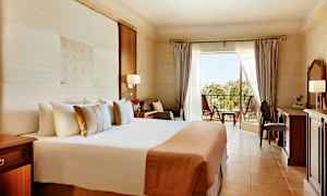 Ett hotellrum med stor säng, skrivbord och balkong med utsikt över grönska. (AI-genererad text)