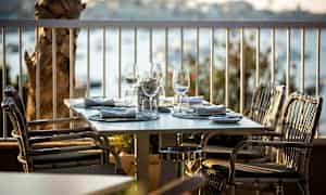 Ett dukat bord med glas och servetter på en utomhusterrass, omgiven av en utsikt över vattnet. (AI-genererad text)