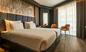 Ett hotellrum med två enkelsängar, en stor fönsteryta med gardiner, två oranga stolar och ett bord. (AI-genererad text)