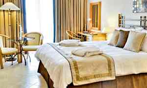 Ett hotellrum med en stor säng, sängkläder och kuddar. Två fåtöljer och ett bord står vid sidan, och en arbetsplats med spegel och skrivbord syns i bakgrunden. (AI-genererad text)