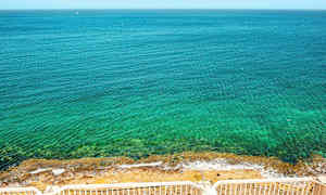 En vy över ett klart blått hav med en stenig strand, sedd från en plats med vita räcken. (AI-genererad text)