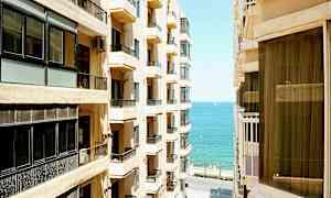 Bild av en stad med höga byggnader på båda sidor av en smal gata, med havet synligt i bakgrunden. (AI-genererad text)