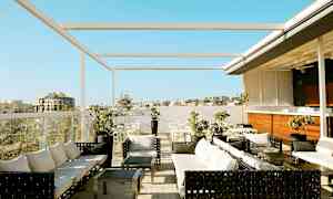 Bild av en takterrass med sittgrupper, bord och parasoll. Utsikt över byggnader och en klarblå himmel. (AI-genererad text)