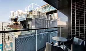 En balkong med modern inredning, inklusive ett runt bord och stolar, med utsikt över en byggnad med glas och metallfasader. (AI-genererad text)