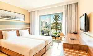 Ett hotellrum med två enkelsängar, ett skrivbord och utsikt över en balkong med pool och träd. (AI-genererad text)