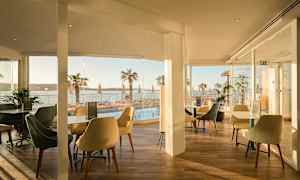 En restaurang med sittplatser och glasväggar med utsikt över en pool och palmer vid havet. (AI-genererad text)