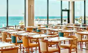 Restaurang med flera bord och stolar, stor fönstervy mot havet. (AI-genererad text)