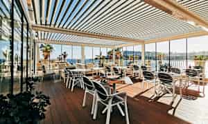 En utomhusterrass med bord och stolar, omgiven av glasväggar och med utsikt över havet och palmer. (AI-genererad text)