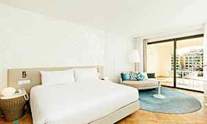 Ett modernt hotellrum med en stor säng, ljusa väggar och elegant inredning. Rummet har en sittgrupp med kuddar samt en rymlig balkong med utsikt. (AI-genererad text)