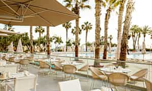 En utomhusrestaurang med flera uppställda bord och stolar, omgärdad av palmer och med en vy över en pool. (AI-genererad text)