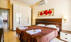 Ett sovrum med två enkelsängar, bruna sängöverkast och vita sängkläder. På väggen ovanför sängarna hänger en tavla med röda blommor. Till vänster finns en garderob med blank vit yta. (AI-genererad text)