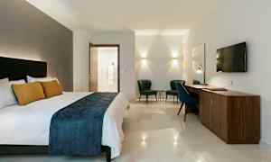 Hotellrum med dubbelsäng, skrivbord, två fåtöljer och väggmonterad TV. (AI-genererad text)