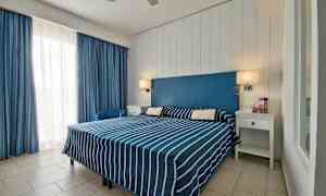 Ett hotellrum med en dubbelsäng klädd i blå- och vitrandiga sängkläder, en blå sänggavel och vita möbler. (AI-genererad text)