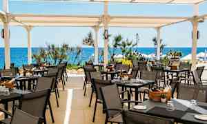 En terrass med uteservering, bord och stolar under ett vitt tak, med havet och vegetation i bakgrunden. (AI-genererad text)