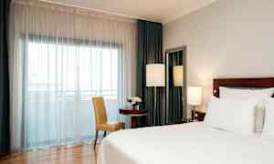 Ett hotellrum med en stor säng, ett bord med en stol, och ett fönster täckt av vita gardiner. (AI-genererad text)