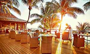 Uteservering med utsikt över havet och solnedgången, palmträd i bakgrunden och en servitör som förbereder ett bord. (AI-genererad text)