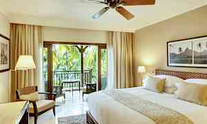 Ett smakfullt inrett hotellrum med en stor säng, sittplats, och balkong som har utsikt över grönskande palmer. (AI-genererad text)