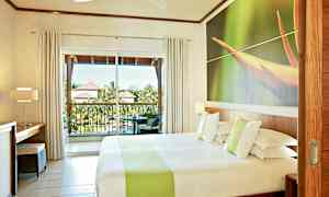 Hotellrum med en dubbelsäng, en stor fönstervägg och en balkong med utsikt över gröna områden och tak av intilliggande byggnader. (AI-genererad text)