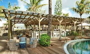 En lounge vid en pool på en tropisk resort, med sittplatser och en bar under träbjälkar. (AI-genererad text)