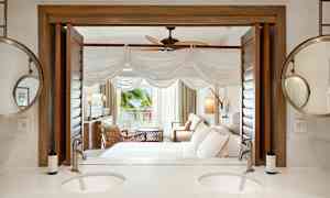 Ett hotellrum med en sittgrupp, säng, och öppning mot en veranda. I förgrunden syns ett badrum med en dubbla handfat och spegel. (AI-genererad text)