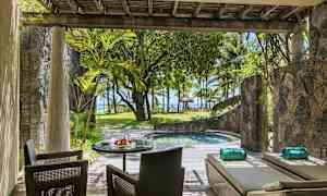 En terrass med sittplatser och solstolar bredvid en liten pool, omgiven av växter och med utsikt mot stranden och palmträd. (AI-genererad text)