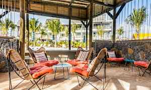 Ett utomhusområde med sittplatser, med korgstolar och färgglada kuddar, samt ett bord på sandgolvet. Trästruktur och gröna växter syns i bakgrunden. (AI-genererad text)