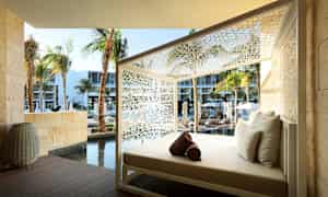 En vilosäng med vita kuddar placerad utomhus, omgiven av vit dekorativ konstruktion. I bakgrunden syns palmer, en pool och moderna byggnader. (AI-genererad text)