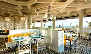 En modern restaurang med geometriskt mönstrat golv, ljusa möbler och stora panoramafönster med utsikt mot havet. (AI-genererad text)