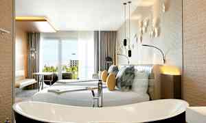 Ett modernt hotellrum med en stor dubbelsäng, dekorativa kuddar och väggdekorationer. Det finns ett fristående badkar och möbler såsom bord och stolar nära en glasdörr med utsikt. (AI-genererad text)