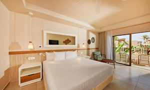 Ett hotellrum med en stor säng, nattduksbord, och en fåtölj med bord. En glasdörr leder ut till en balkong med utsikt över ett grönskande område. (AI-genererad text)