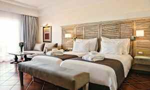 Ett hotellrum med två separata sängar, en soffa, ett bord med en vas blommor och ett fönster med gardiner. (AI-genererad text)