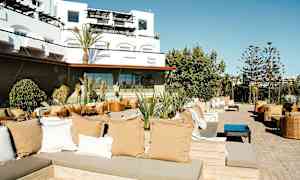 En utomhus lounge område med sittgrupper, kuddar och växter, med en modern byggnad i bakgrunden. (AI-genererad text)