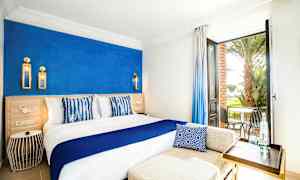 Ett ljust hotellrum med en blå accentvägg, en stor säng med vit och blå inredning, samt en öppen dörr med utsikt över en terrass med stolar och palmer. (AI-genererad text)