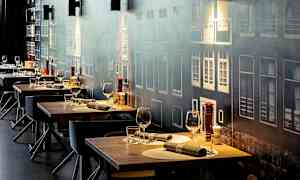 Ett antal dukade restaurangbord med glas och bestick, mot en vägg med fasadmotiv. (AI-genererad text)