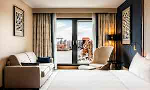 Ett hotellrum med en säng, en soffa och en stol framför ett stort fönster med utsikt över en stad och vatten. (AI-genererad text)