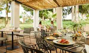 En utomhusrestaurang med dukade bord och gröna växter i bakgrunden. (AI-genererad text)