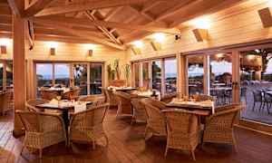 Inomhusmiljö med matbord och stolar av rotting, stora fönster med utsikt mot en terrass och landskap utanför. (AI-genererad text)