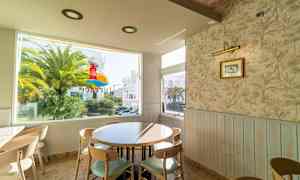 Ett café med ett rundt bord och stolar nära ett fönster som visar en palm och en byggnad utanför. (AI-genererad text)