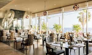 En restaurang med moderna bord och stolar, stora fönster som visar utsikt över havet och palmträd. (AI-genererad text)