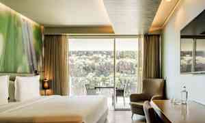 Ett hotellrum med en dubbelsäng, sittgrupp, ett bord med stolar samt en stor glasdörr som öppnar upp till en balkong med utsikt över natur och klippor. (AI-genererad text)