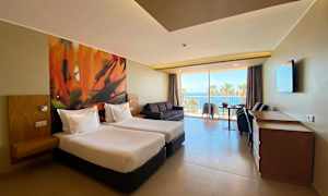 Hotellrum med två enkelsängar, soffa, bord och stolar. Fönster med utsikt mot palmer och hav. (AI-genererad text)