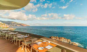 Restaurangterrass med bord och stolar, med utsikt över havet och en klippig kustlinje under en klar himmel. (AI-genererad text)